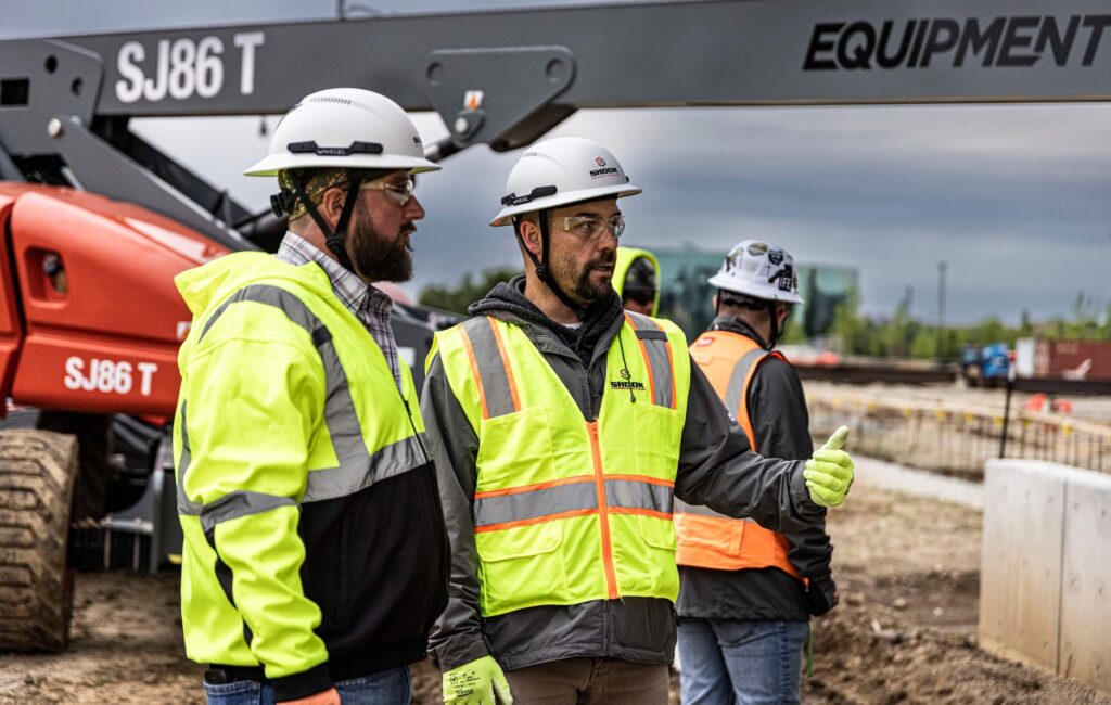 Two construction workers in safety vests and helmets talk on a worksite, while two others work in the background near equipment.
