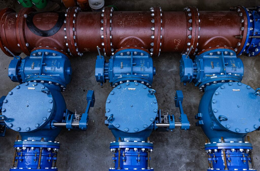 Three large blue industrial valves are connected to a horizontal brown pipeline. The equipment is placed on a concrete floor and the scene shows a close-up, top-down view of the mechanical setup.