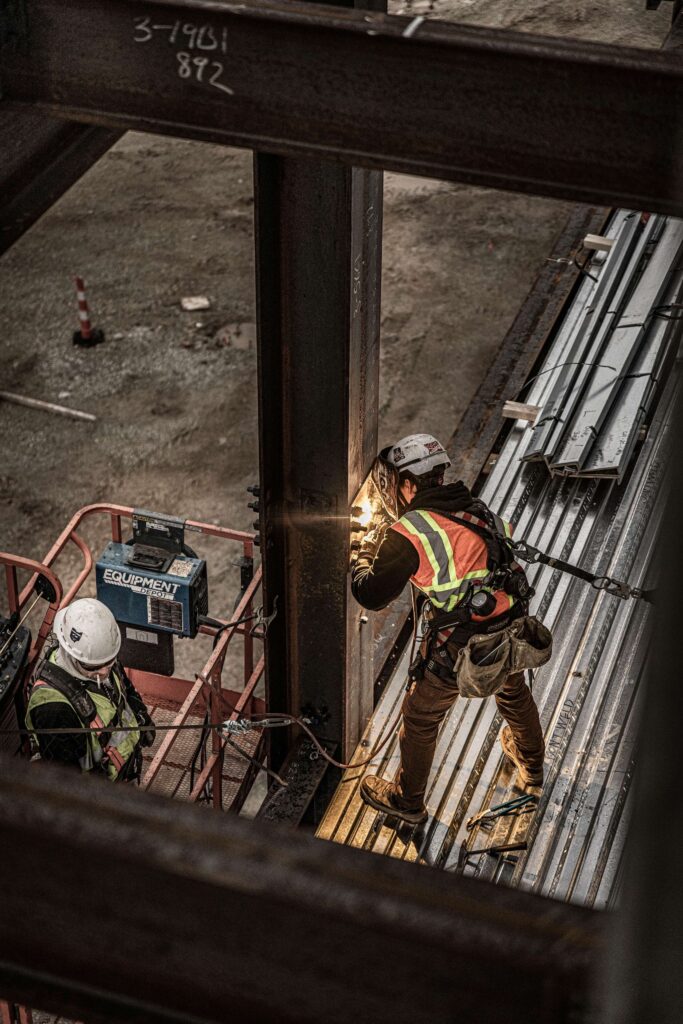 Two construction workers in safety gear work on a steel structure. One operates equipment, causing sparks, while the other stands nearby. Both wear helmets and reflective vests at a construction site.