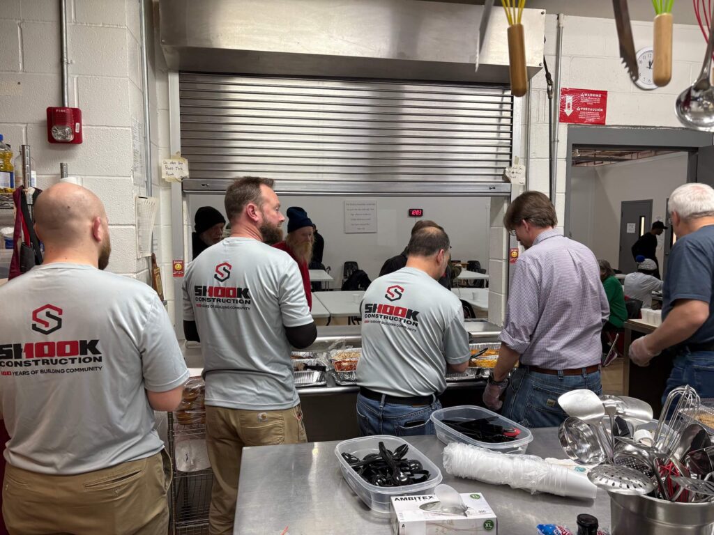 Volunteers in gray Shook Construction shirts serve food from a kitchen counter to people waiting on the other side. The kitchen is busy with various utensils and supplies on the counter.