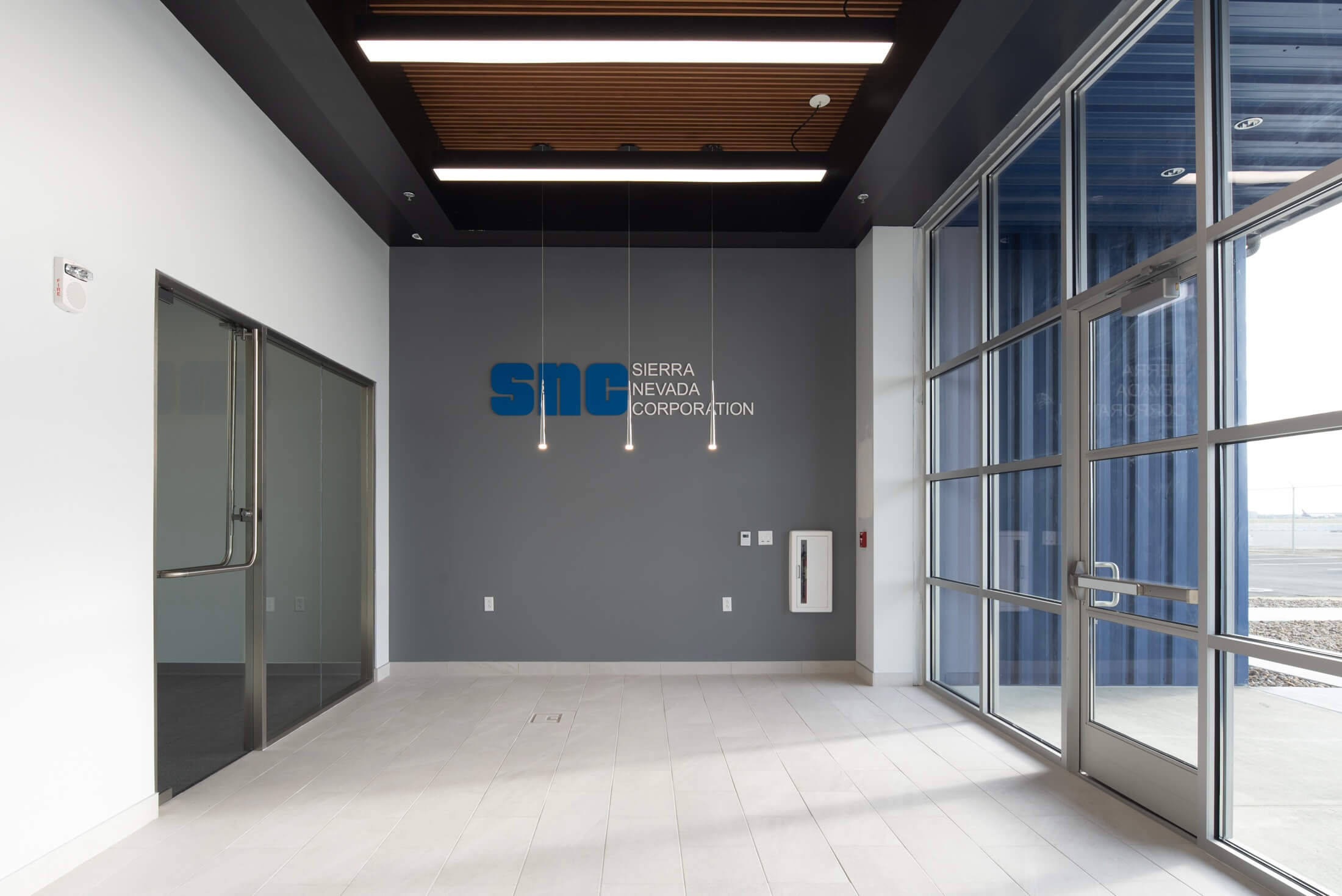 Modern office lobby with glass doors, gray walls, white floor tiles, hanging pendant lights, and Sierra Nevada Corporation logo on the wall. Large windows provide natural light from the right side.