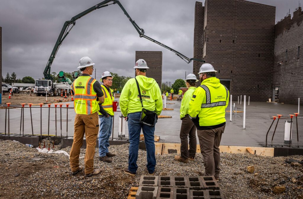 Five construction workers in safety gear and hard hats stand on a gravel area at a building site, discussing plans. Behind them, concrete walls and construction equipment are visible under a cloudy sky.