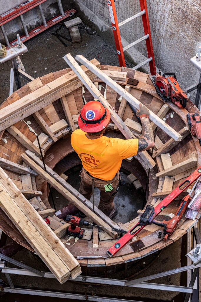 Construction worker in a yellow shirt and orange hard hat stands inside a circular wooden structure surrounded by tools, wooden planks, and red ladders, as seen from above.