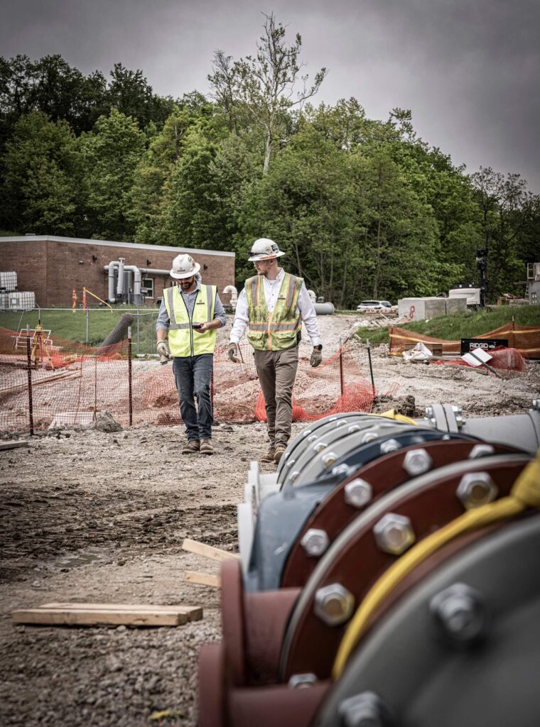 Two construction workers wearing safety vests and helmets walk at a worksite with large pipes in the foreground, orange fencing, trees, and a brick building in the background under a cloudy sky.