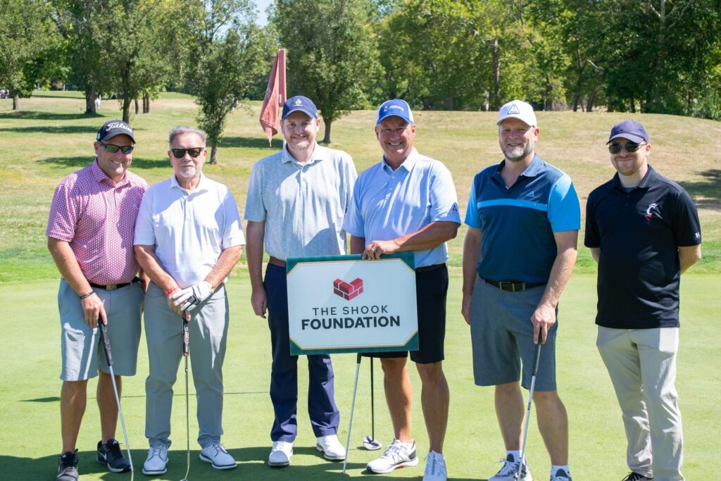 Six men stand on a golf course holding clubs, smiling next to a sign that reads The Shook Foundation. Trees and grass are visible in the background under a sunny sky.