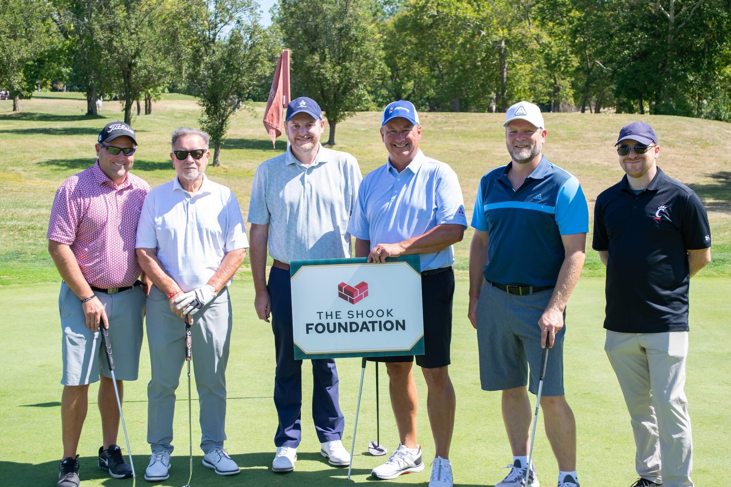 Six men stand on a golf course holding clubs, smiling next to a sign that reads The Shook Foundation. Trees and grass are visible in the background under a sunny sky.