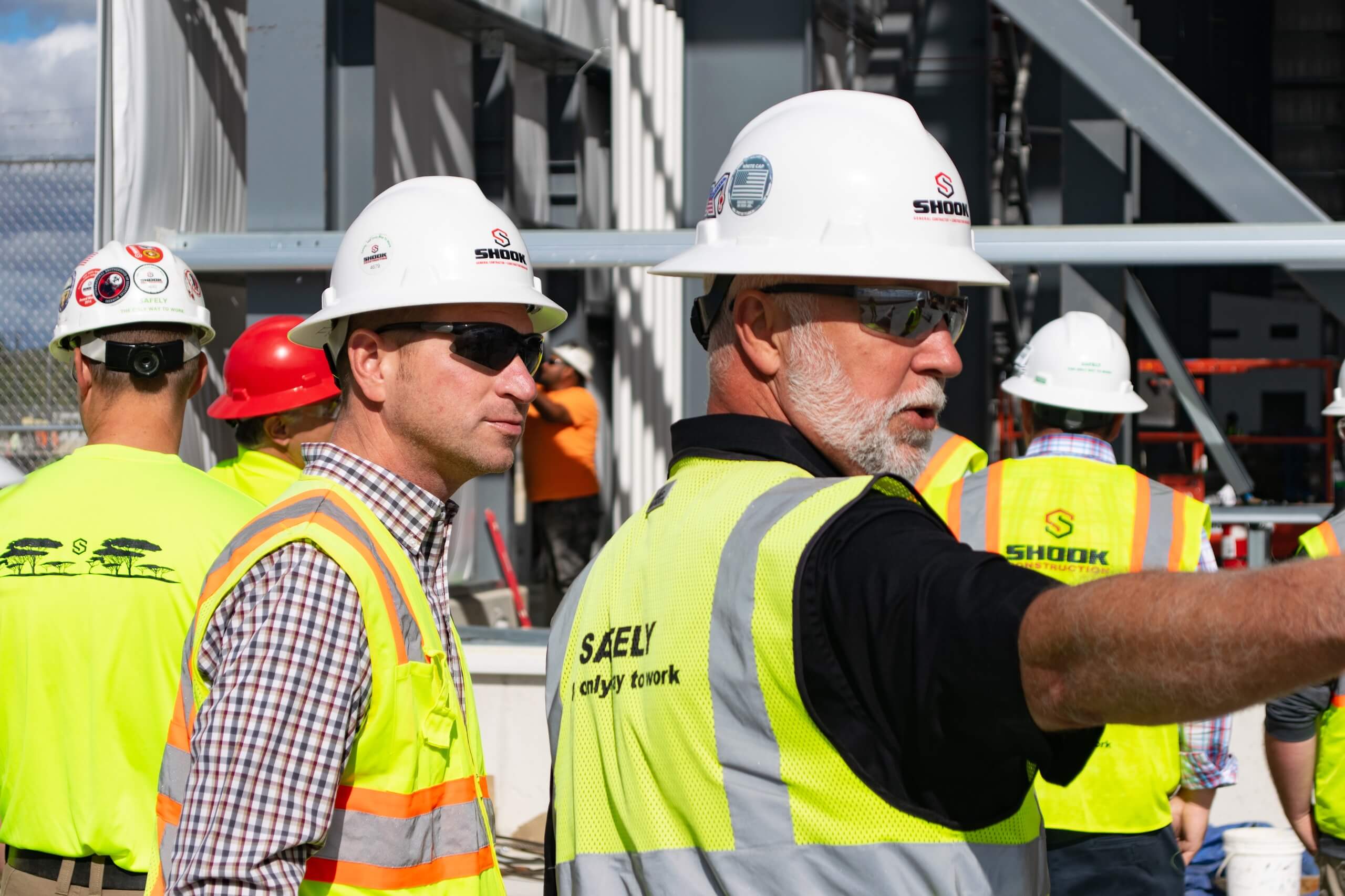 Two construction workers wearing hard hats and safety vests discuss a project on site. One points ahead while others work in the background, surrounded by structural steel beams in sunlight.