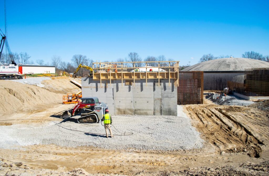 A construction worker stands near machinery by a large concrete wall at a construction site, surrounded by dirt, equipment, and partially built structures under a clear blue sky.