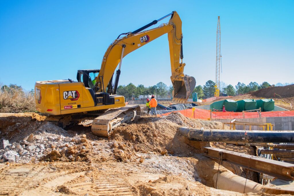 A construction site with a yellow excavator moving dirt, two workers in safety vests nearby, and cranes, pipes, and fencing in the background under a clear blue sky.