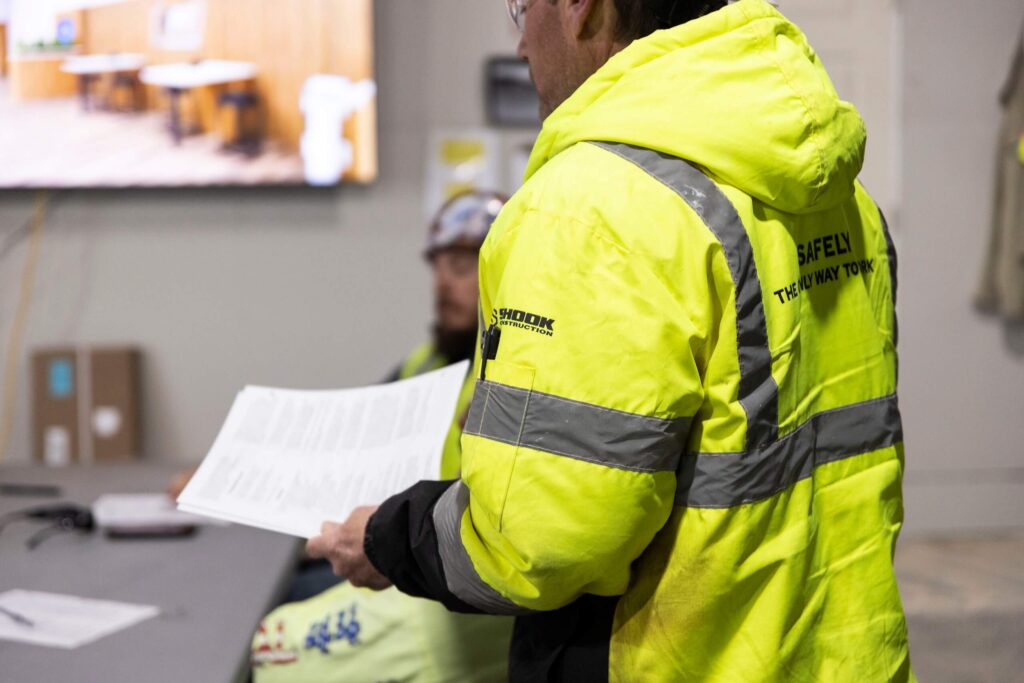 A person in a bright yellow safety jacket holds documents, standing beside another seated person in a similar jacket inside an indoor workspace.
