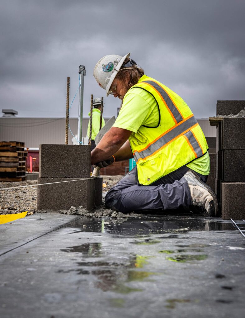 A construction worker wearing a hard hat and yellow safety vest kneels on the ground, laying cement blocks at an outdoor work site under a cloudy sky.