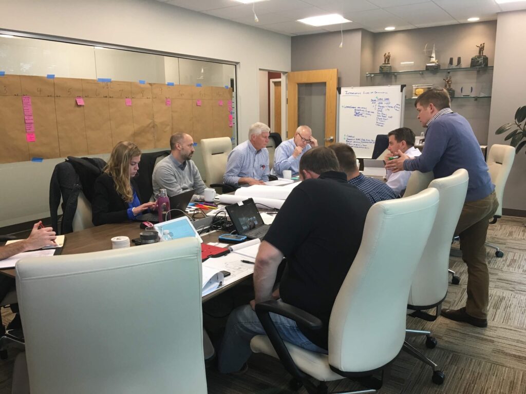 Eight people sit around a conference table with laptops, papers, and notes. One person stands and speaks to the group. A whiteboard and a large board with sticky notes are visible in the background.