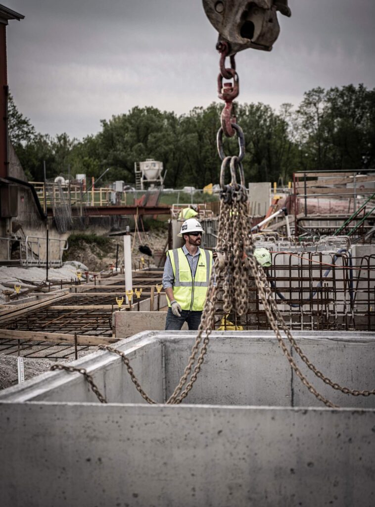 A construction worker in a safety vest and helmet stands near a concrete slab being lifted by chains on a busy construction site, with trees and equipment in the background.