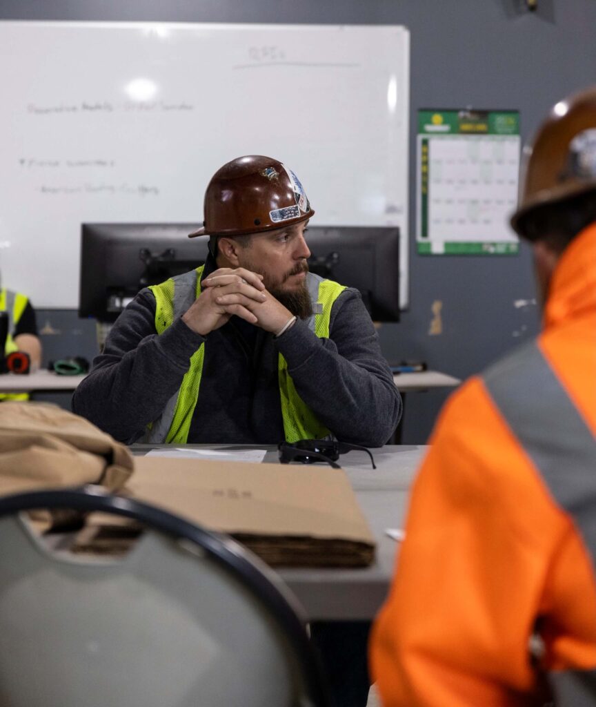 A man in a hard hat and reflective vest sits at a table, looking to the side. He is in an office with a whiteboard, computer monitors, and another person in a hard hat and orange jacket nearby.