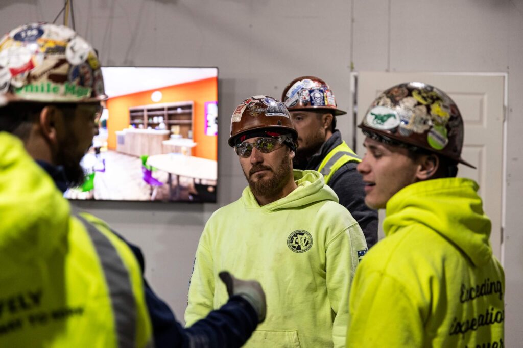 Four construction workers wearing hard hats and safety gear stand indoors, having a discussion. One worker gestures while others listen. A wall-mounted screen in the background displays a colorful room image.