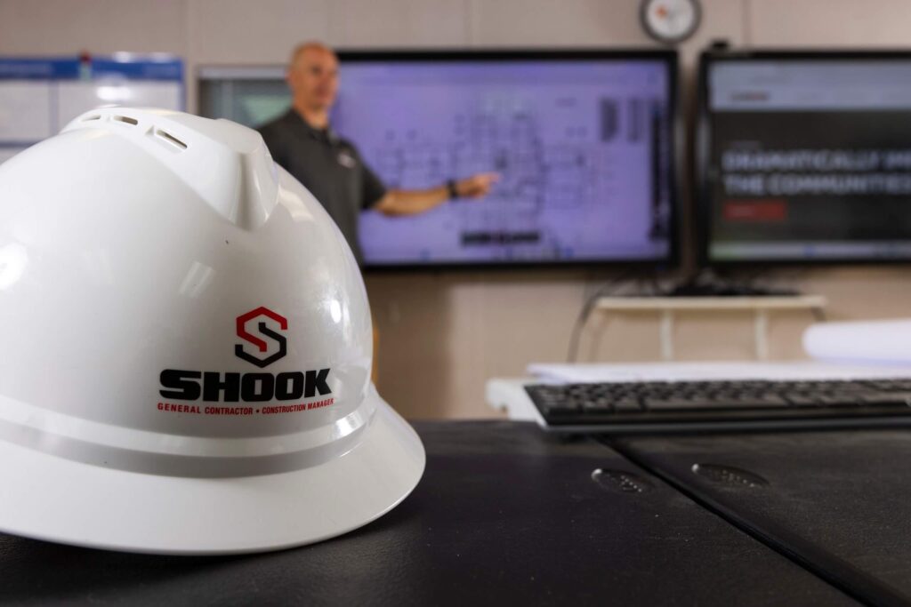 A white hard hat with the Shook logo sits on a table in the foreground. In the background, a person points at a large screen displaying building plans.