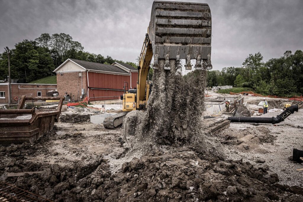 A construction site with an excavator dumping gravel onto the ground, surrounded by dirt, debris, and nearby buildings on a cloudy day.
