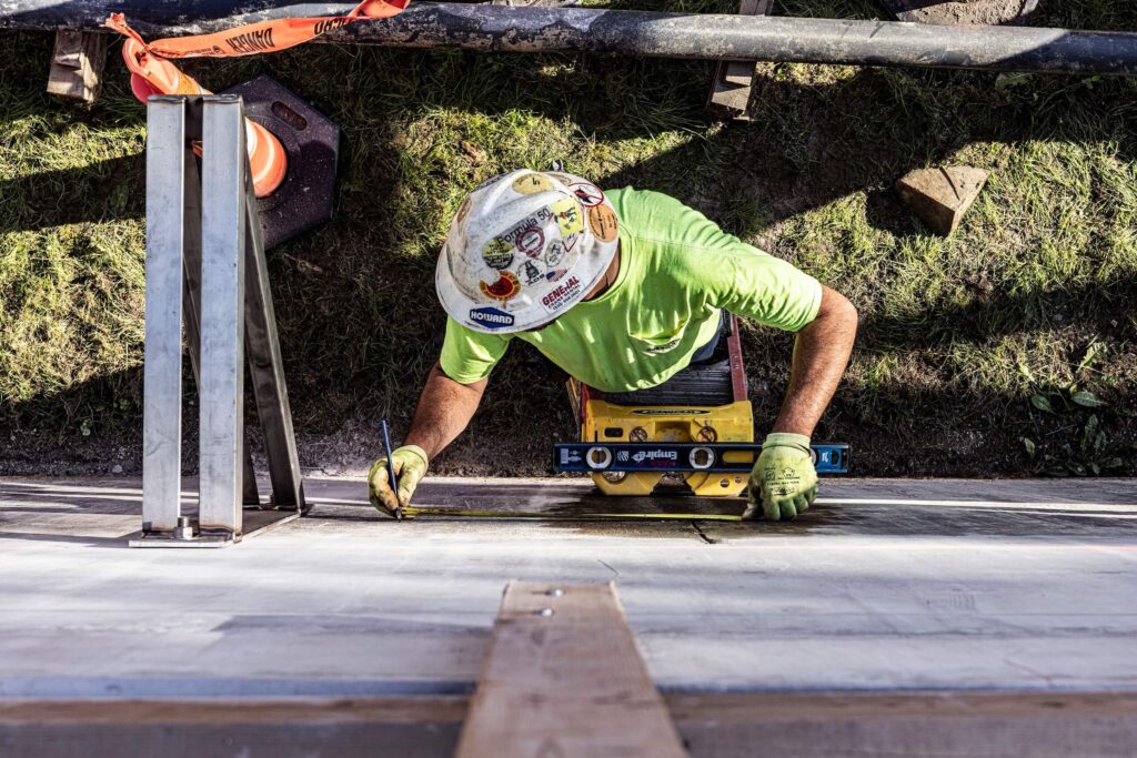A construction worker in a bright shirt and hard hat measures a concrete wall with a level while standing on a ladder, viewed from above. Grass and safety equipment are visible nearby.