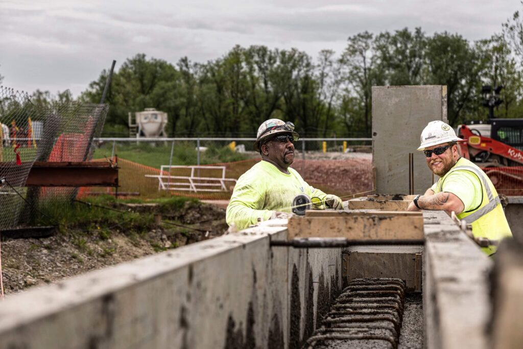 Two construction workers in safety gear stand at a concrete formwork, smiling and working together on a construction site with trees and equipment in the background.