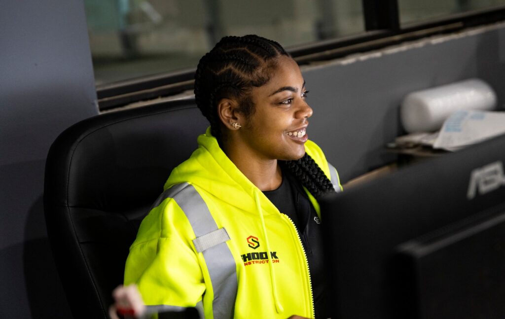 A woman wearing a bright yellow reflective jacket sits at a desk, smiling while looking at a monitor. She has braided hair and is indoors, with office supplies visible around her.