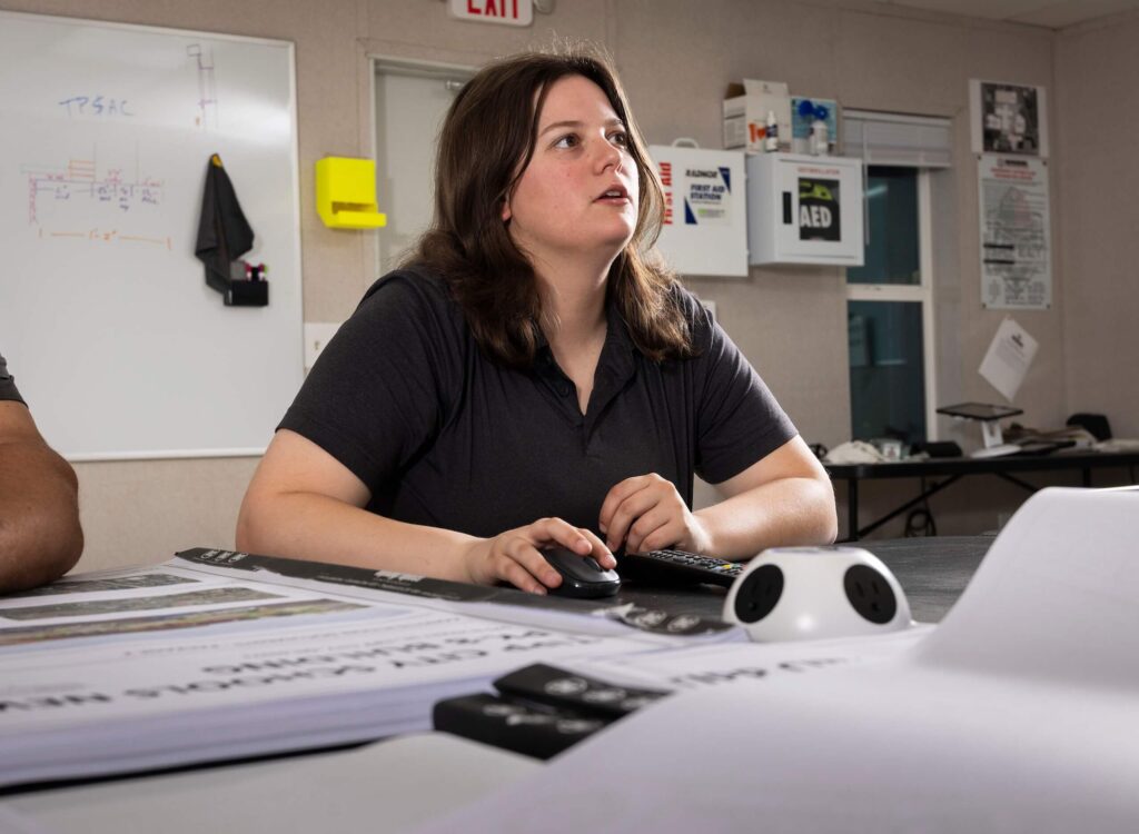 A person with shoulder-length brown hair sits at a table using a computer mouse, looking up attentively. Papers and office supplies are on the table; a whiteboard and safety equipment are visible in the background.