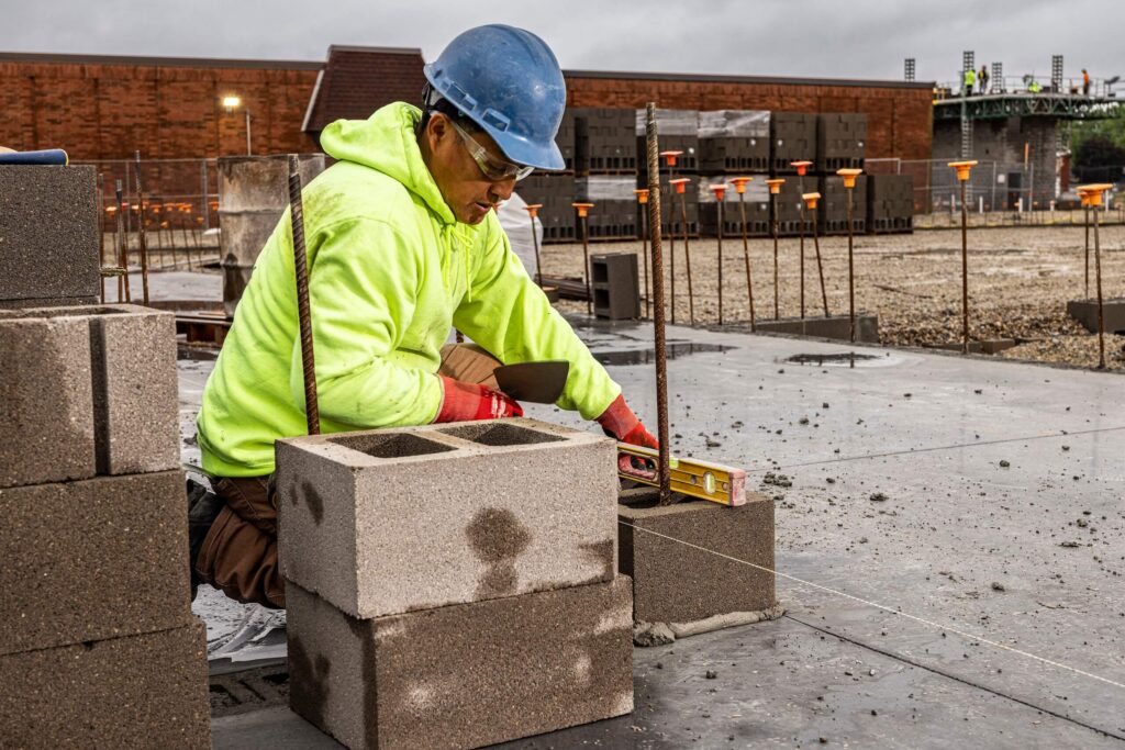 A construction worker wearing a blue helmet and yellow safety hoodie uses a level to place concrete blocks at a building site.