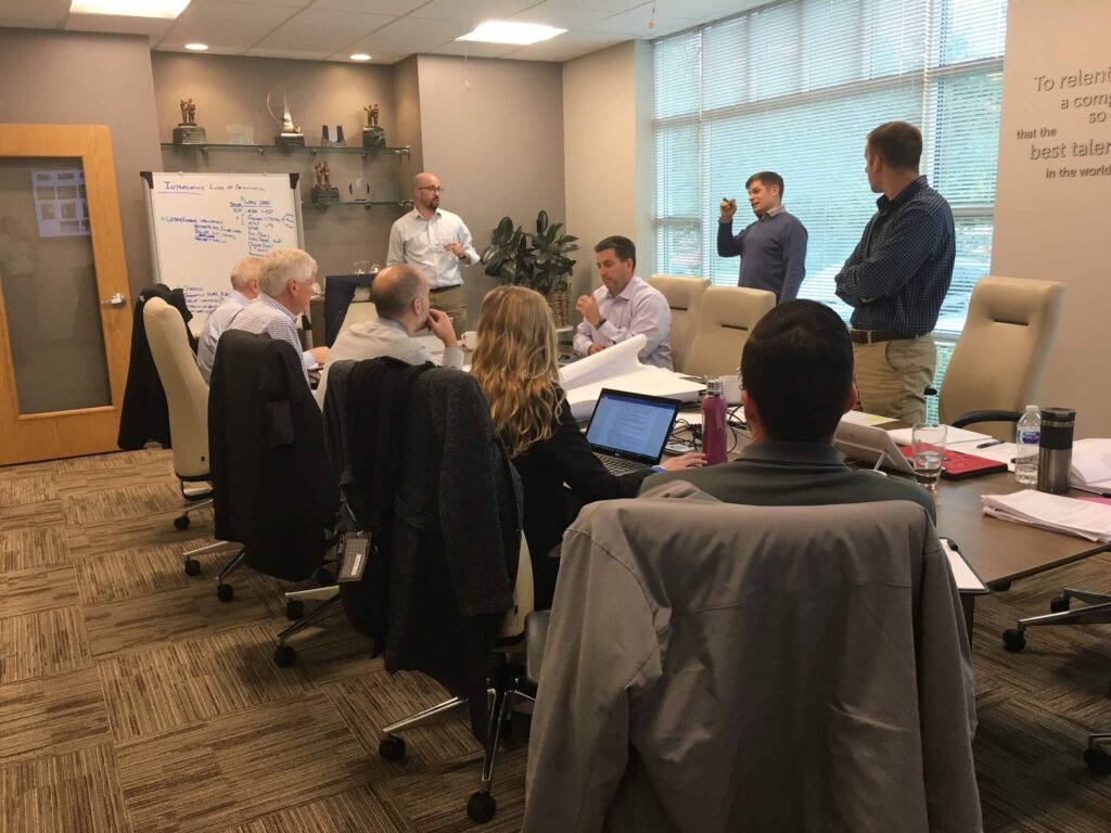 A group of people have a meeting in a conference room. Some are seated around a table with laptops and papers, while others stand near a whiteboard covered in writing. Large windows let in natural light.