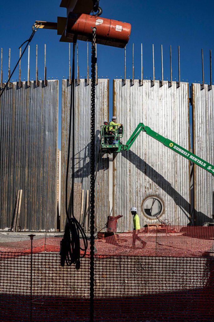Two construction workers operate a green lift in front of a tall concrete wall, while another worker walks below. Orange safety netting and construction equipment are visible under a clear blue sky.
