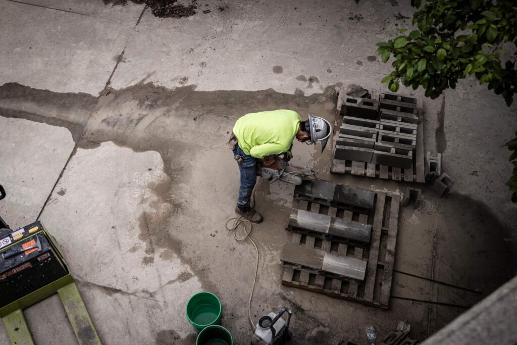 A construction worker in a yellow shirt and helmet uses a power saw to cut concrete blocks near stacked pallets. Buckets and tools are nearby on a dusty outdoor surface.
