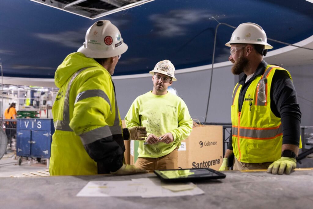 Three construction workers wearing hard hats and safety vests stand indoors, engaged in conversation. Construction materials, documents, and a tablet are visible on the table in front of them.
