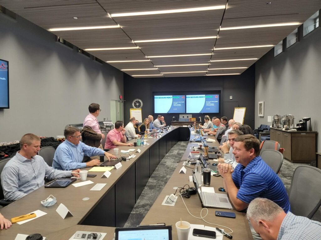 A group of people are seated around a large conference table in a modern meeting room, engaged in discussion. Laptops, papers, and drinks are on the table, and presentation screens are visible at the front of the room.