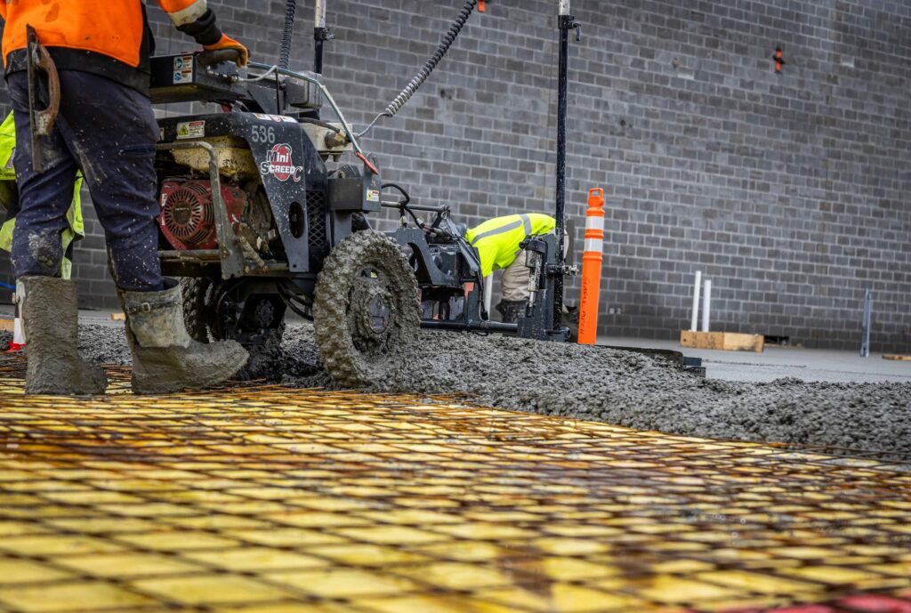 Construction workers pour and spread wet concrete over a rebar grid at a building site. One worker operates machinery while another, wearing a high-visibility jacket, bends down by a concrete barrier wall.
