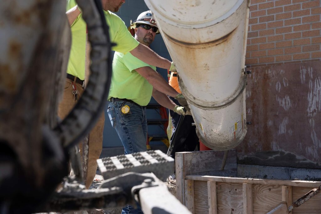 Two construction workers in yellow shirts guide a large concrete chute to pour cement into a wooden form next to a brick building.