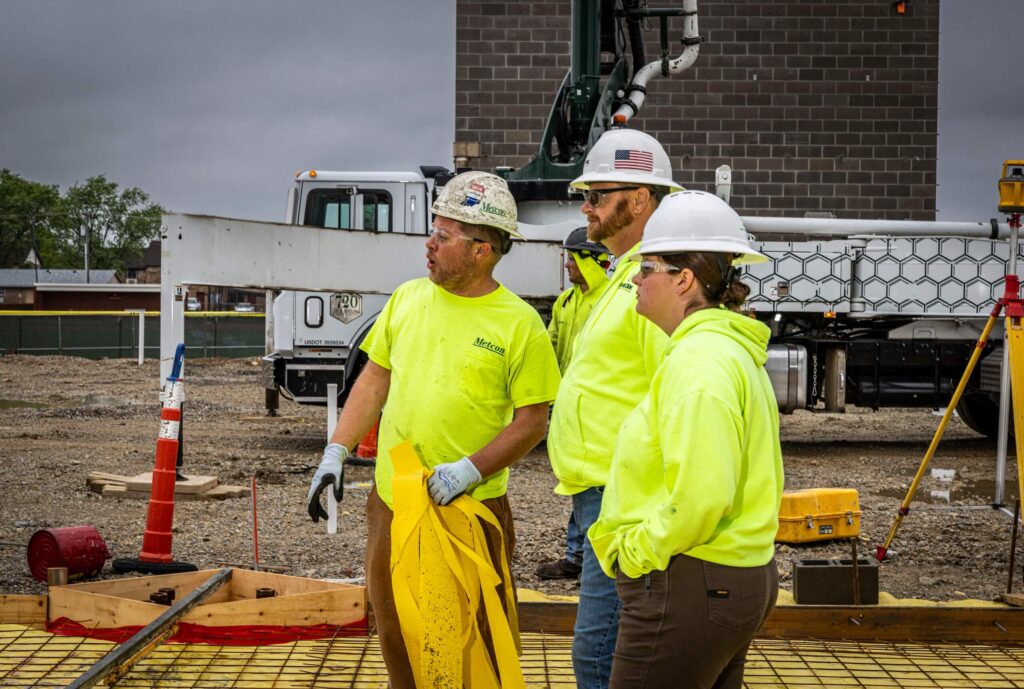 Three construction workers in hard hats and neon safety jackets stand on a building site, talking and looking in the same direction. Construction equipment and a brick building are visible in the background.