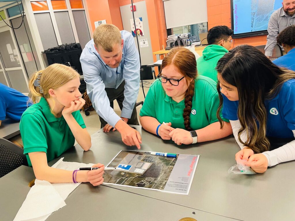 A teacher leans over a table, pointing to a paper as three students watch and listen attentively during a classroom activity.
