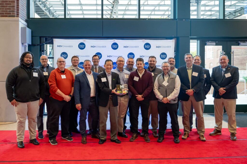 A group of people pose indoors on a red carpet in front of an Indy Chamber backdrop. The person in the center holds a small trophy or award. Everyone is dressed in business or business-casual attire.
