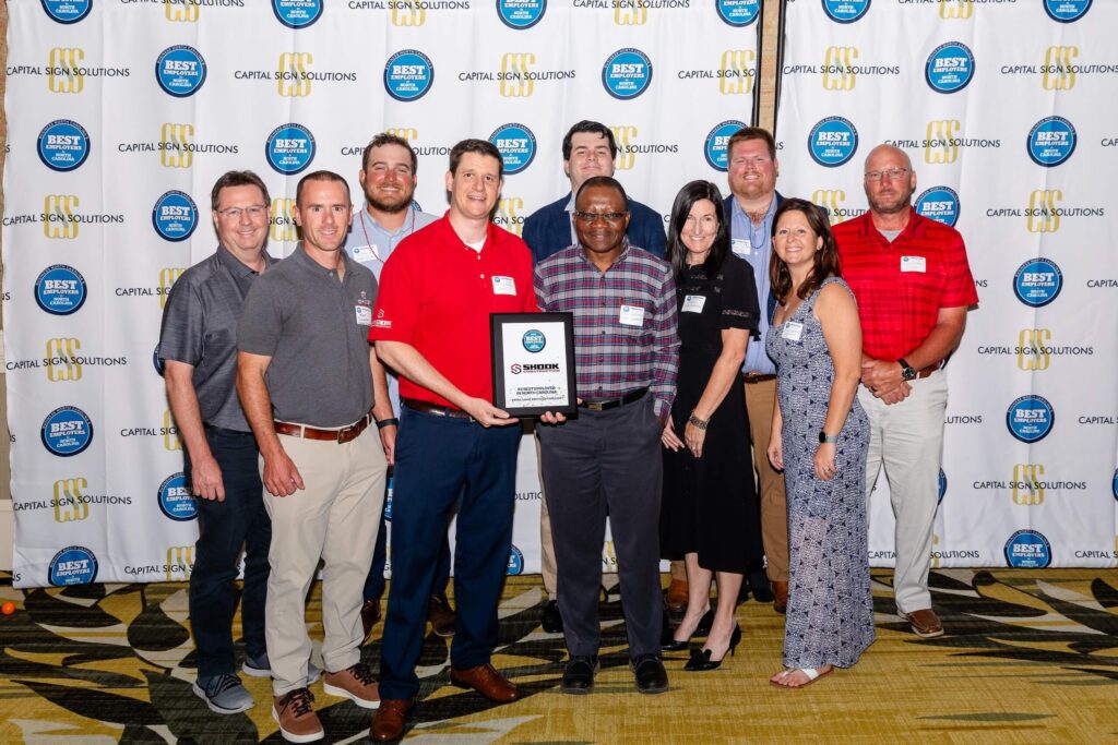 A group of eleven people pose indoors, with one person holding a framed award. They stand on a patterned floor in front of a backdrop featuring circular Best logos and text.
