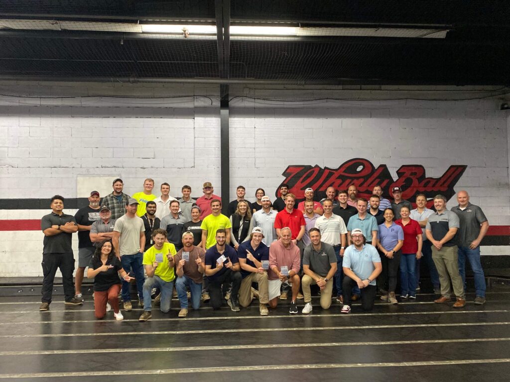 A large group of people pose together inside a sports facility with Whirly Ball written on the wall behind them. Most are smiling, some crouch in front, and others stand in rows.