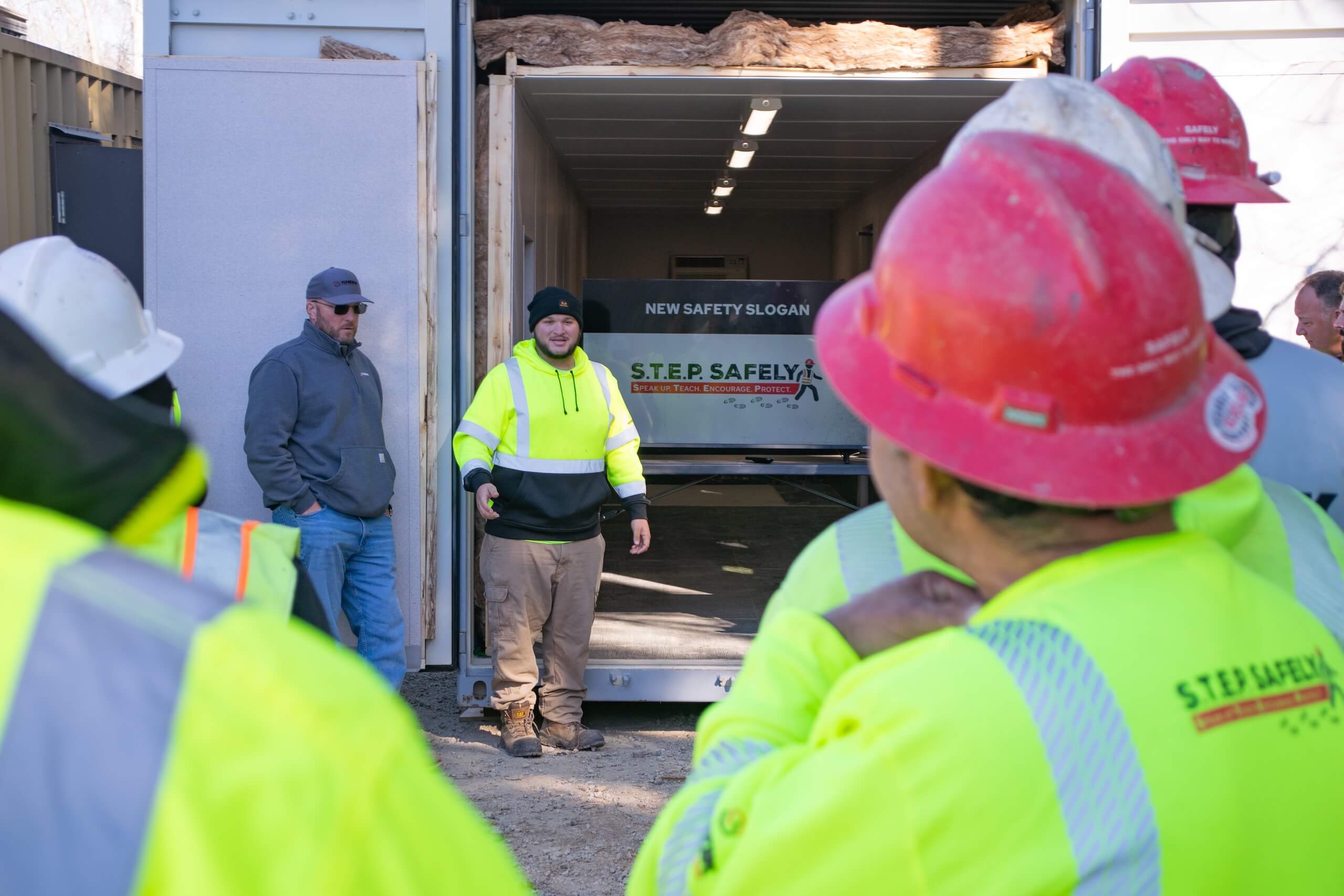 A group of construction workers in safety gear listens to a man in a bright yellow jacket speaking outside a shipping container displaying a STEP SAFELY safety slogan inside.
