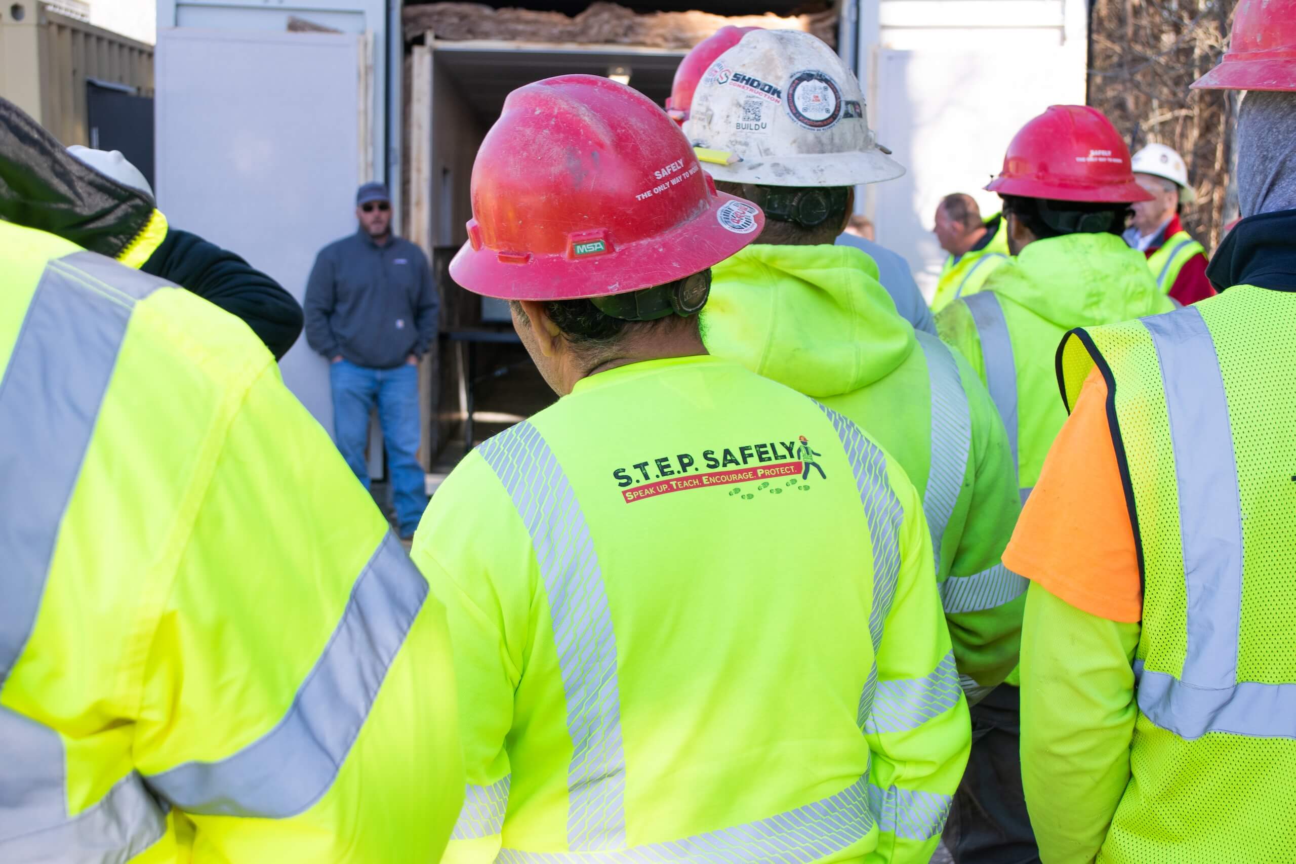 Construction workers in high-visibility jackets and hard hats gather outdoors. The back of one workers shirt reads S.T.E.P. SAFELY. A person in a gray hoodie stands near an open shipping container.