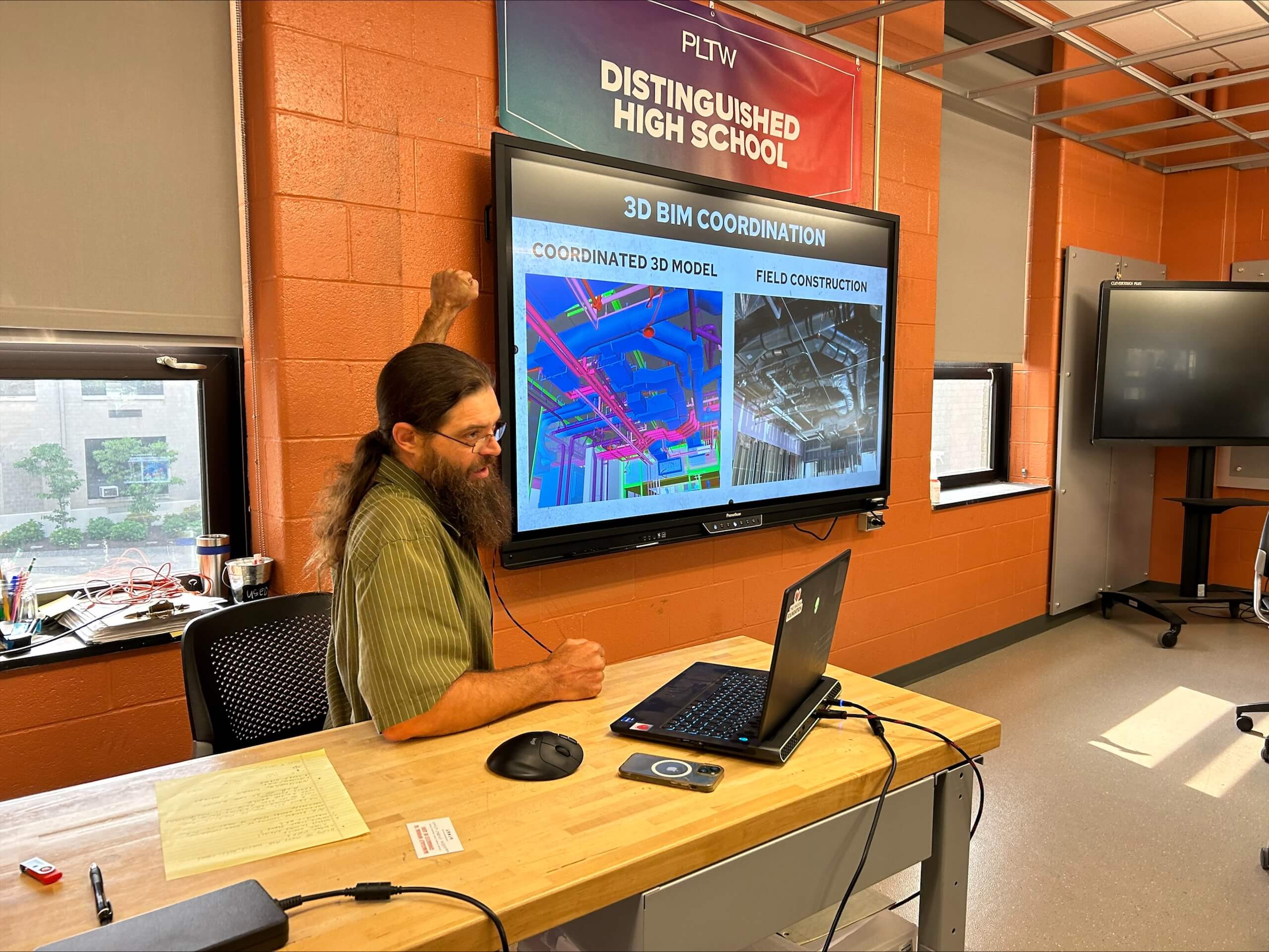 A man with long hair and a beard gestures at a screen showing 3D BIM coordination images in a classroom. A laptop sits on the desk, and a Distinguished High School sign is above the monitor.