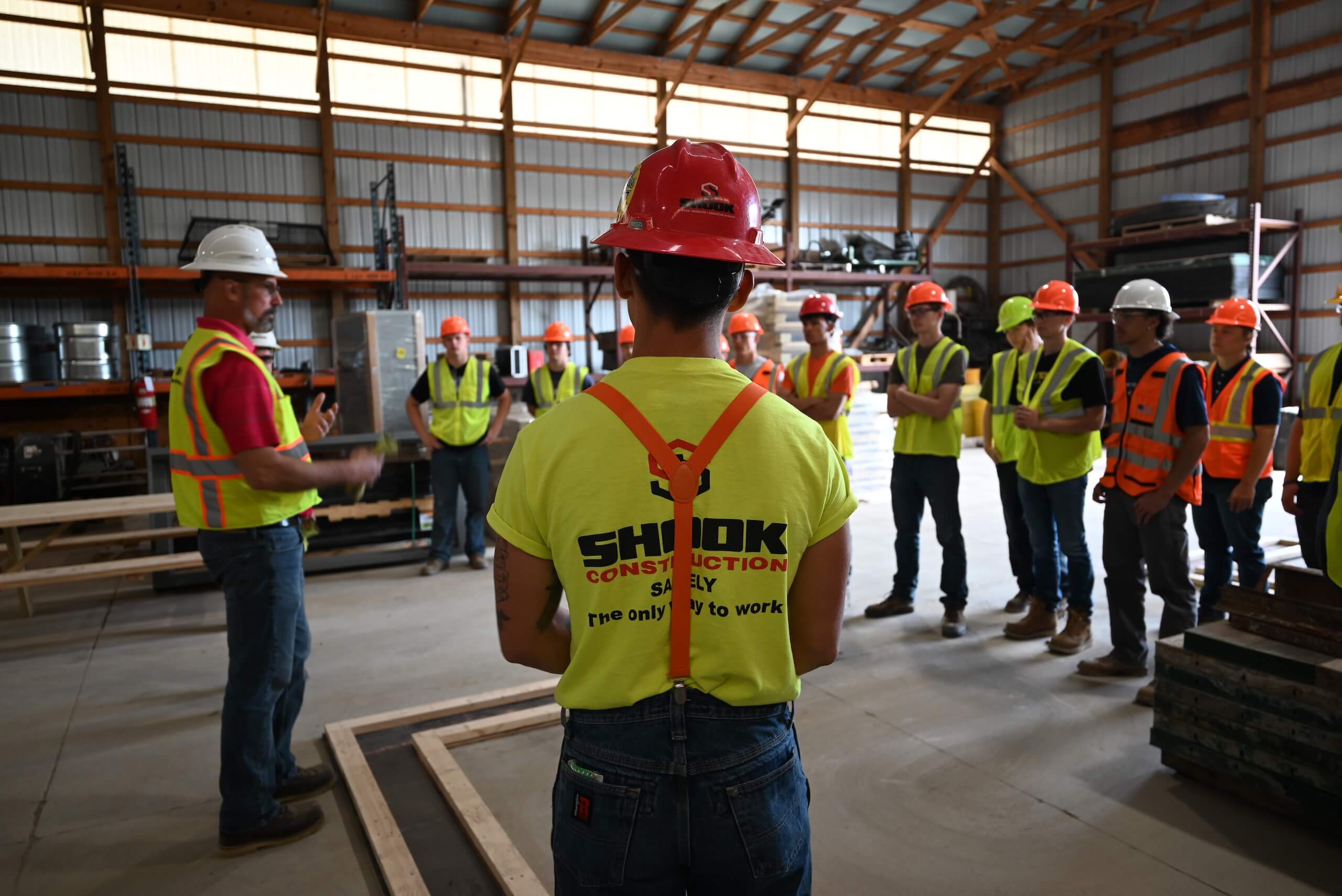 A group of construction workers in safety gear stand in a warehouse, listening to a person speaking at the front. Most wear yellow vests and hard hats; one in the foreground wears a shirt reading SHIOK CONSTRUCTION..