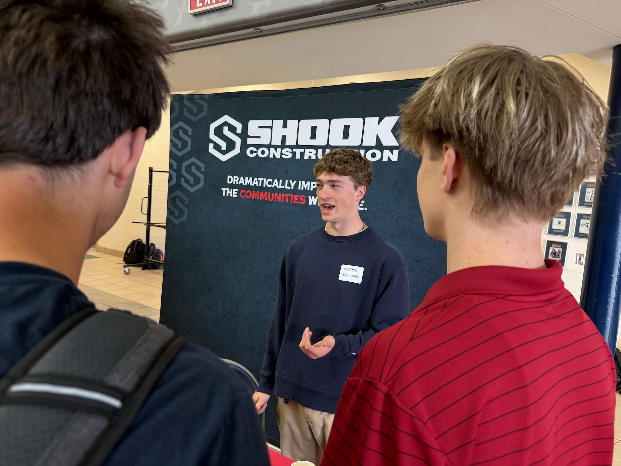 A young man speaks to two others at a table in front of a Shook Construction sign at an indoor event. The sign reads Dramatically IMPROVING the COMMUNITIES we SERVE..