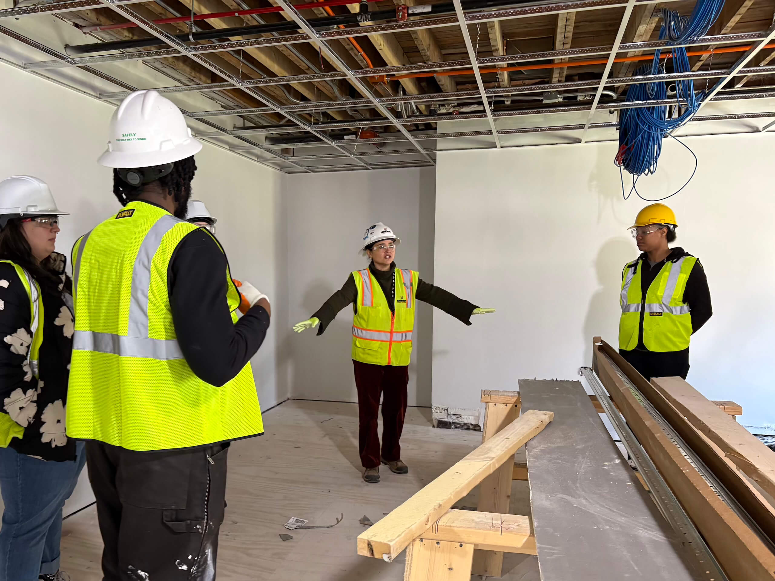 Five people wearing hard hats and safety vests stand in a partially constructed room, discussing plans. Exposed ceiling wiring and construction materials are visible around them.