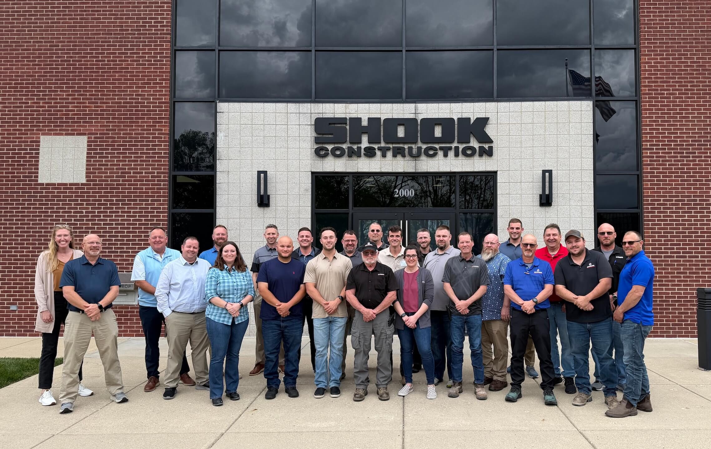 A group of 23 people stands smiling in front of a building with a sign that reads SHOOK CONSTRUCTION above the entrance. The group appears to be a mix of men and women, dressed in business casual and work attire.