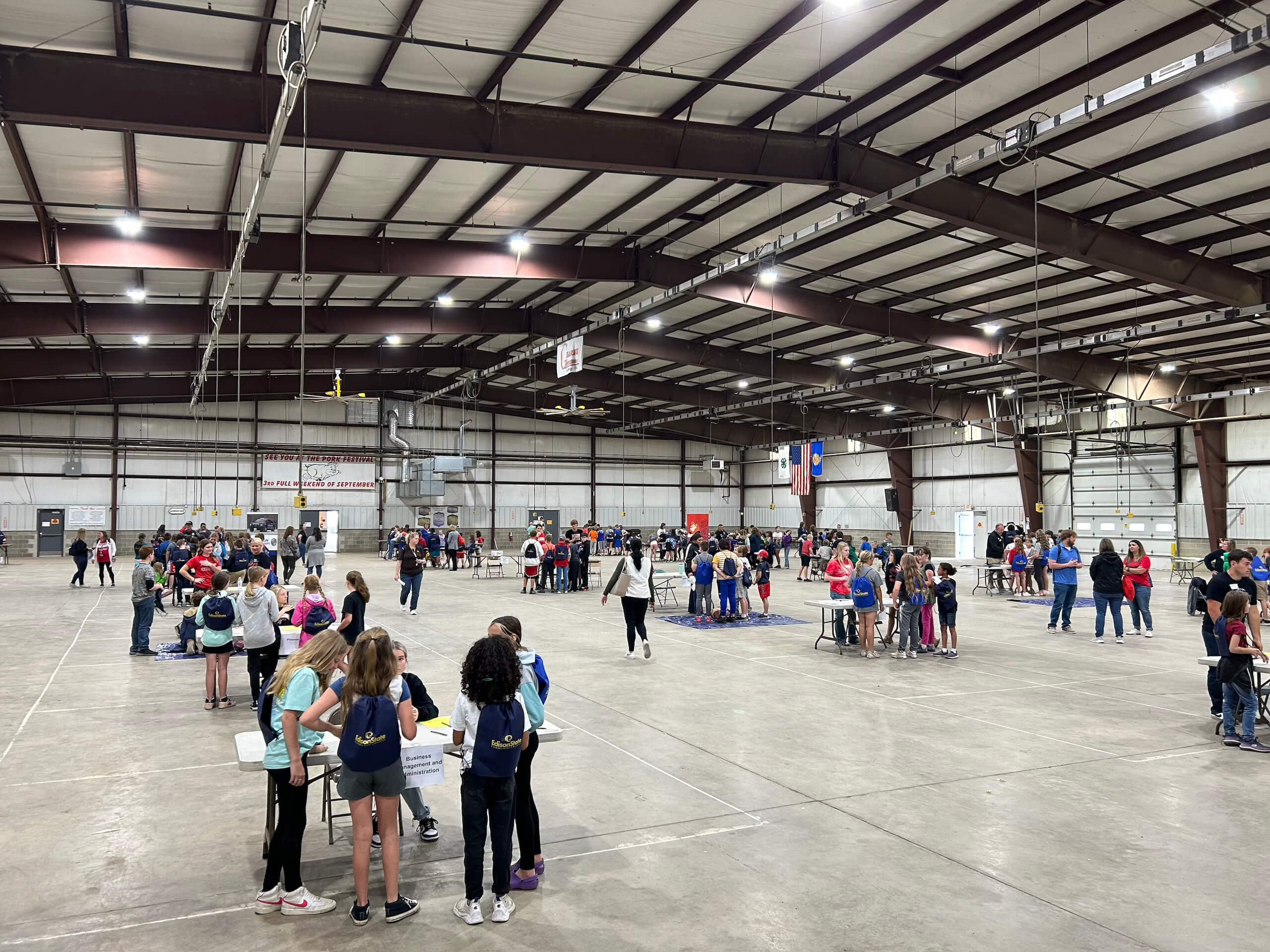 Groups of children and adults gather around tables inside a large, open indoor facility with a high ceiling and exposed beams. The atmosphere appears casual and social.