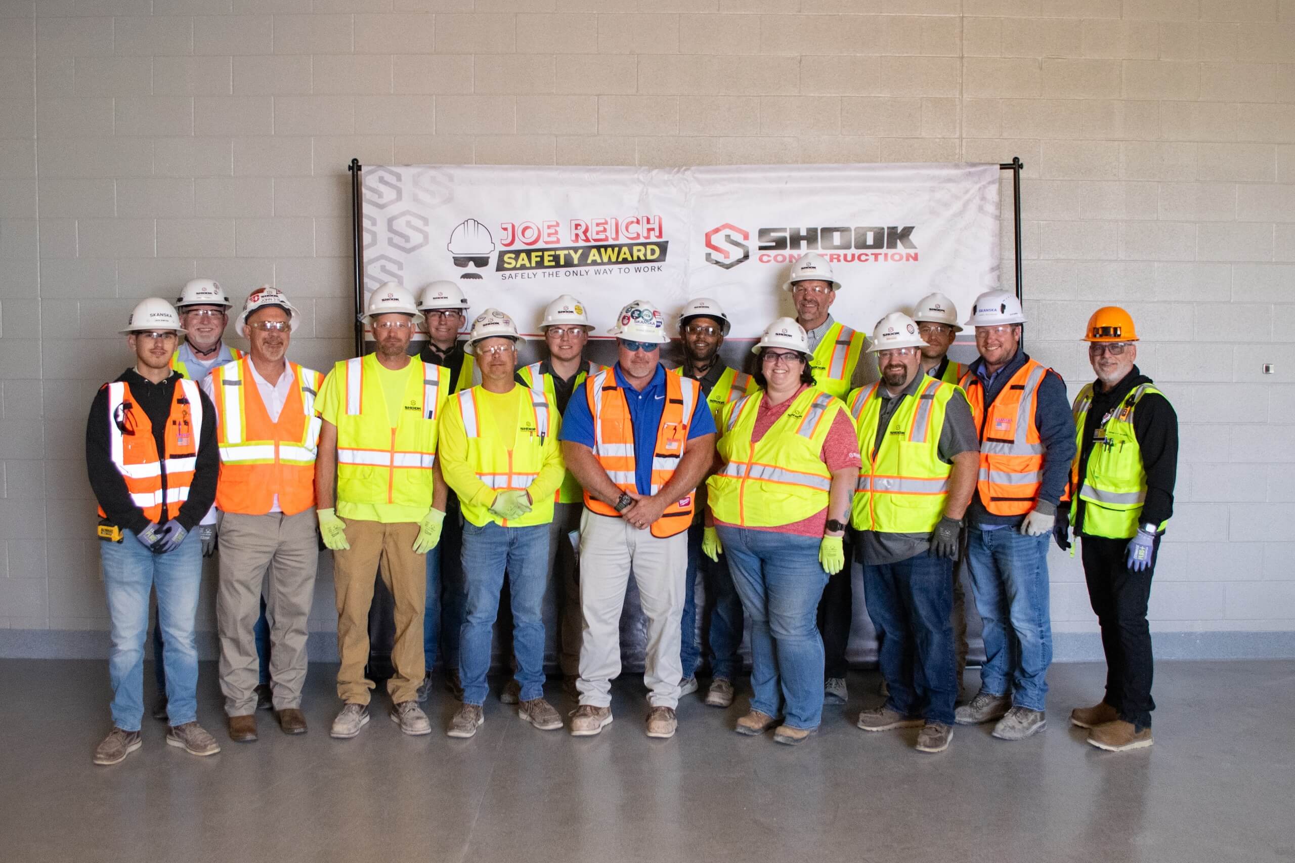 Sixteen construction workers in safety gear pose indoors in front of banners reading Joe Reich Safety Award and Shook Construction..