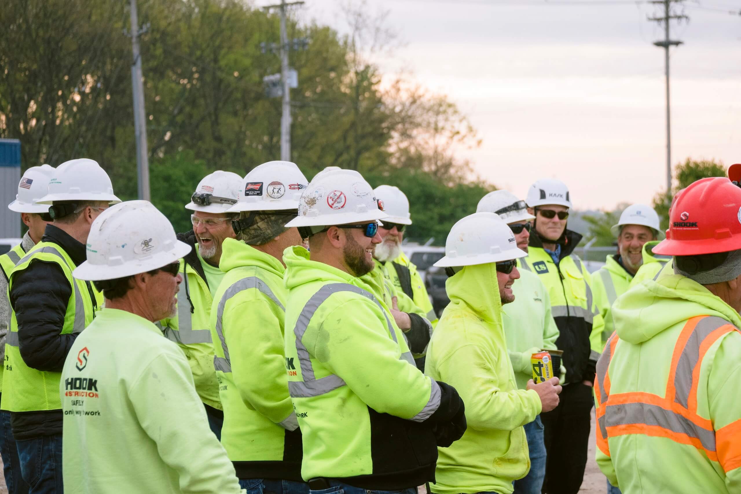 A group of construction workers in safety gear, including hard hats and neon yellow vests, stand together outdoors, talking and smiling. Trees and power lines are visible in the background.