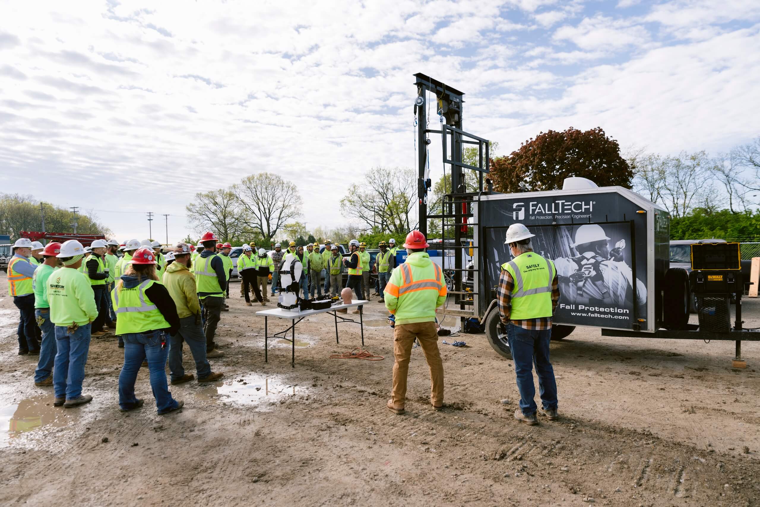 A group of construction workers in safety vests and helmets gather outdoors around a FallTech safety demonstration booth on a dirt lot, listening to a presentation.