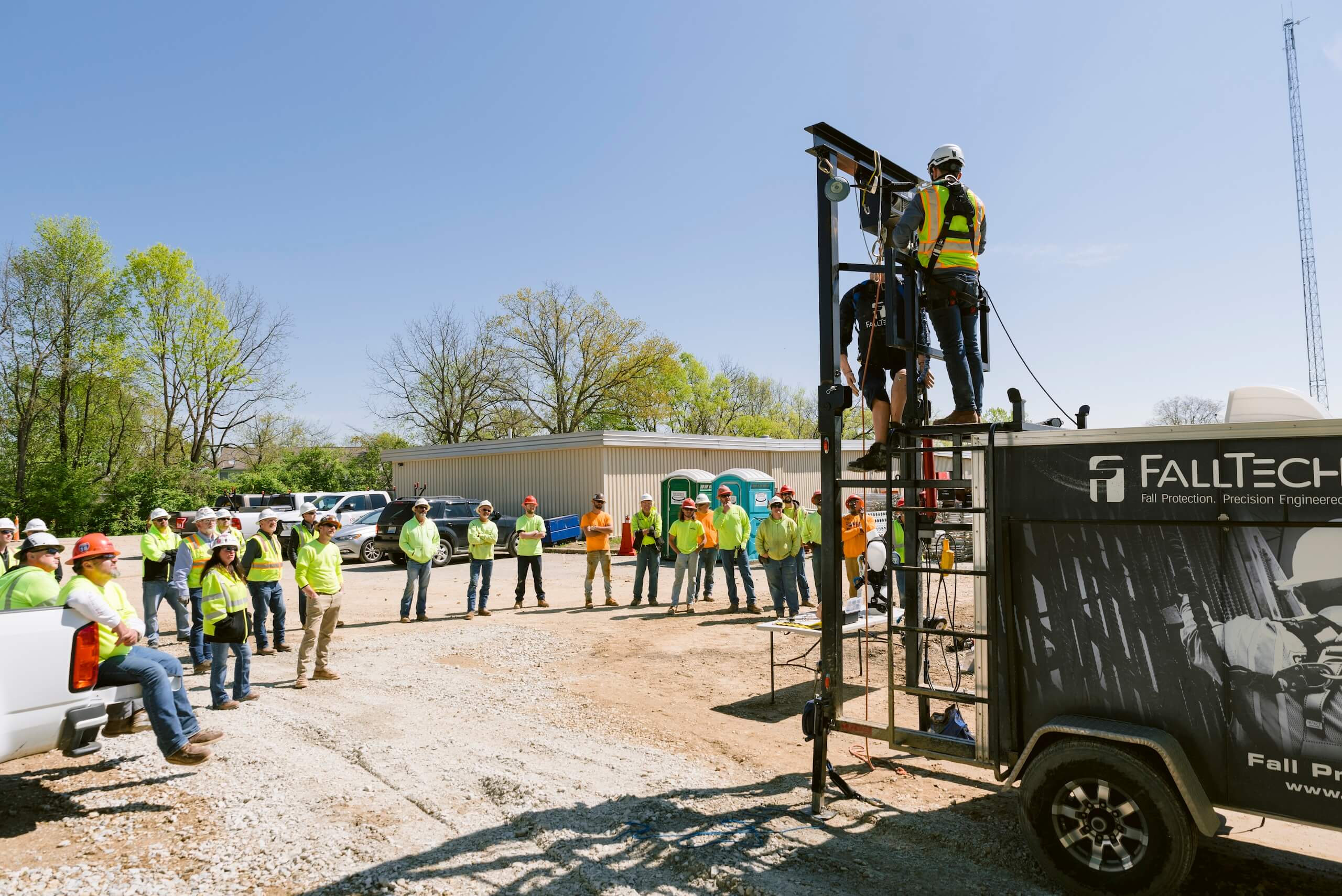 A group of people in safety gear watch a fall protection demonstration outdoors, where two workers use safety harnesses on a tall metal frame next to a trailer labeled FallTech..